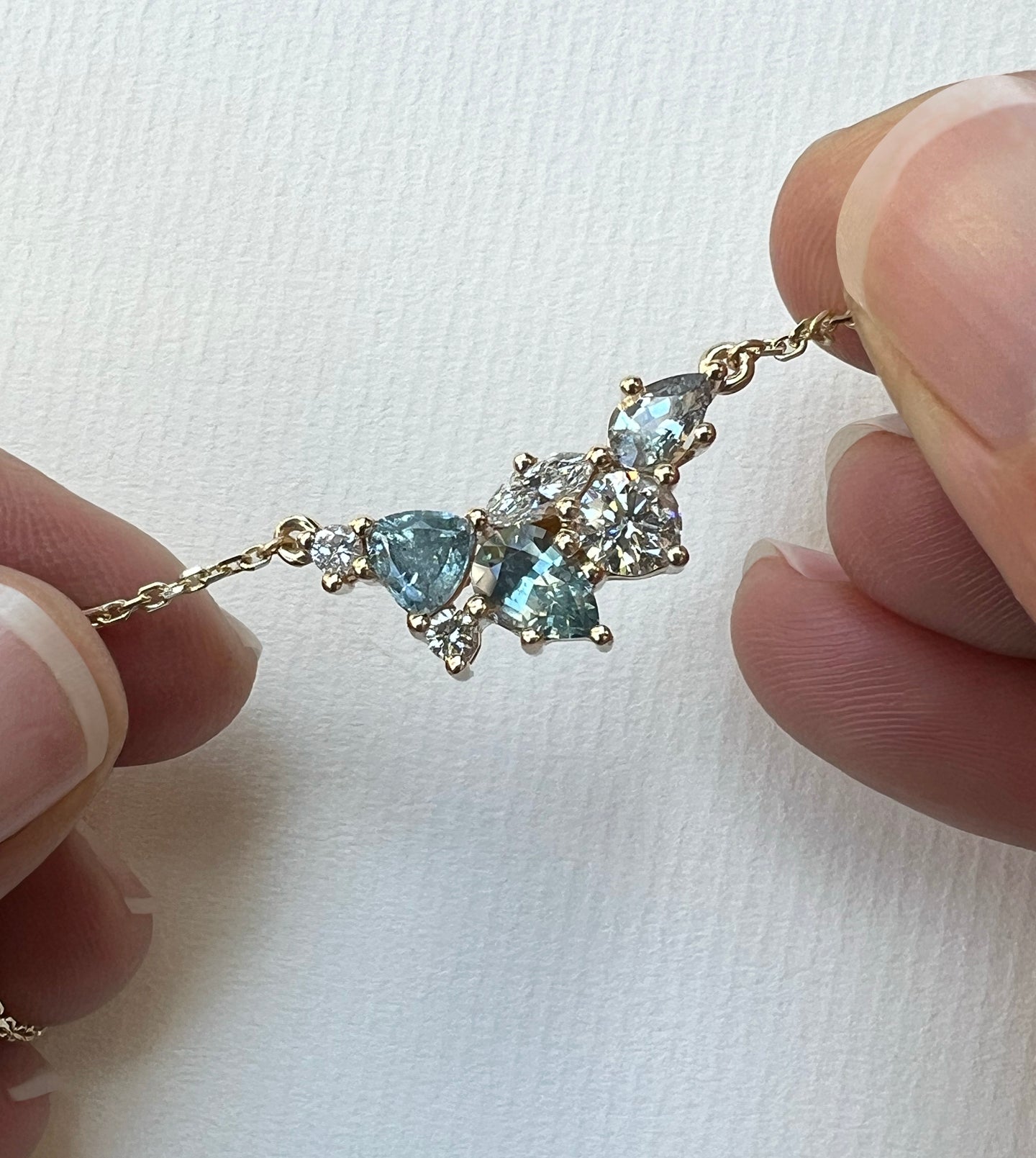Close-up of a 14k gold gemstone cluster necklace with silvery-blue Montana sapphires and diamonds, held between fingers
