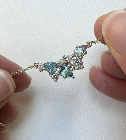 Close-up of a 14k gold gemstone cluster necklace with silvery-blue Montana sapphires and diamonds, held between fingers