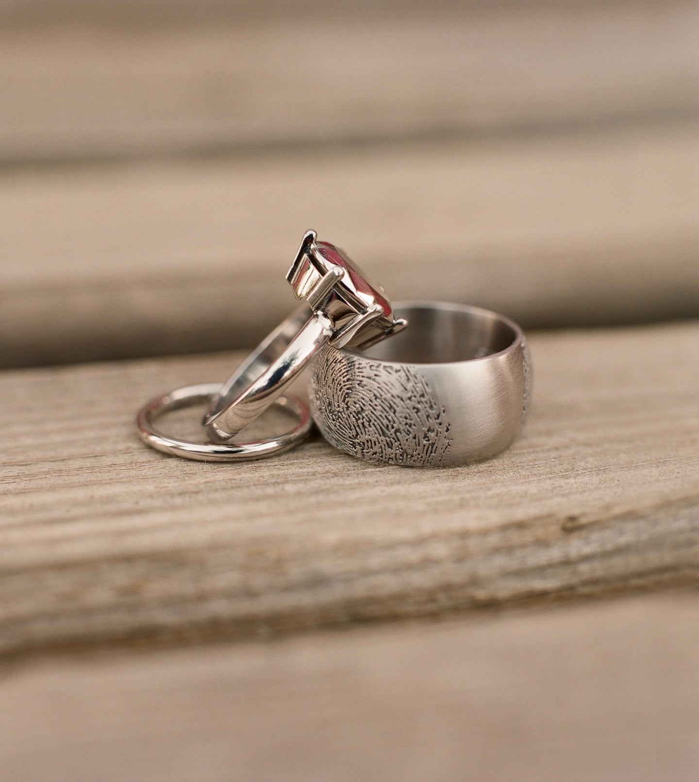 Two palladium wedding rings on weathered wood—a wide brushed band engraved with a fingerprint and a slim ring with a pink faceted gemstone