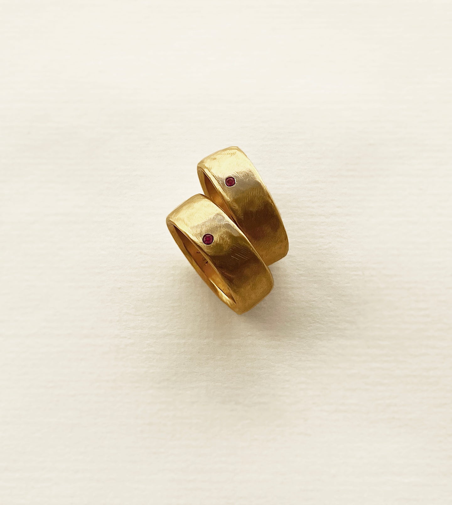 Pair of wide 18k yellow gold wedding bands with hand-carved tool-mark texture, each flush-set with a small ruby