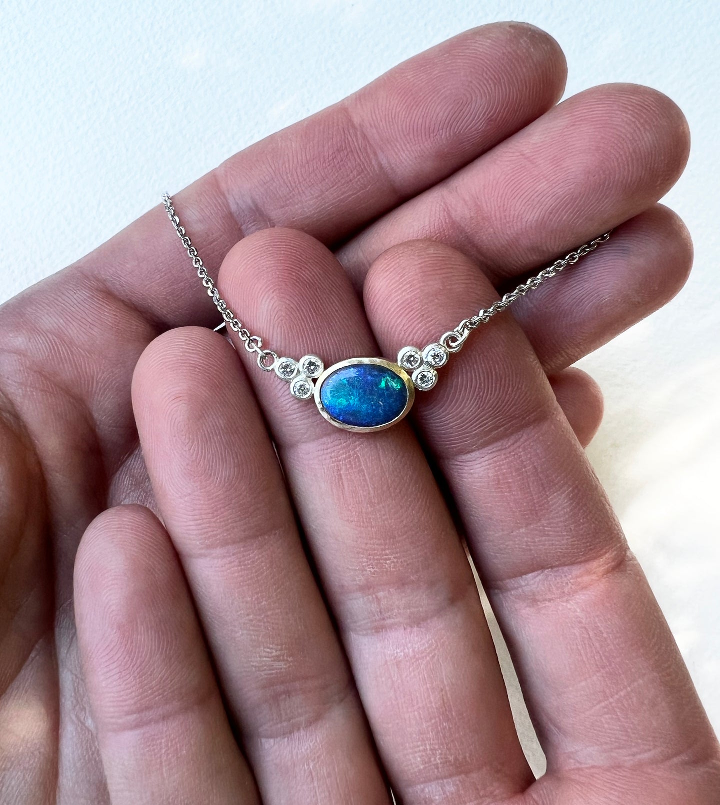 Opal and diamond necklace held in hands, blue-green opal in a hammered gold bezel with diamonds